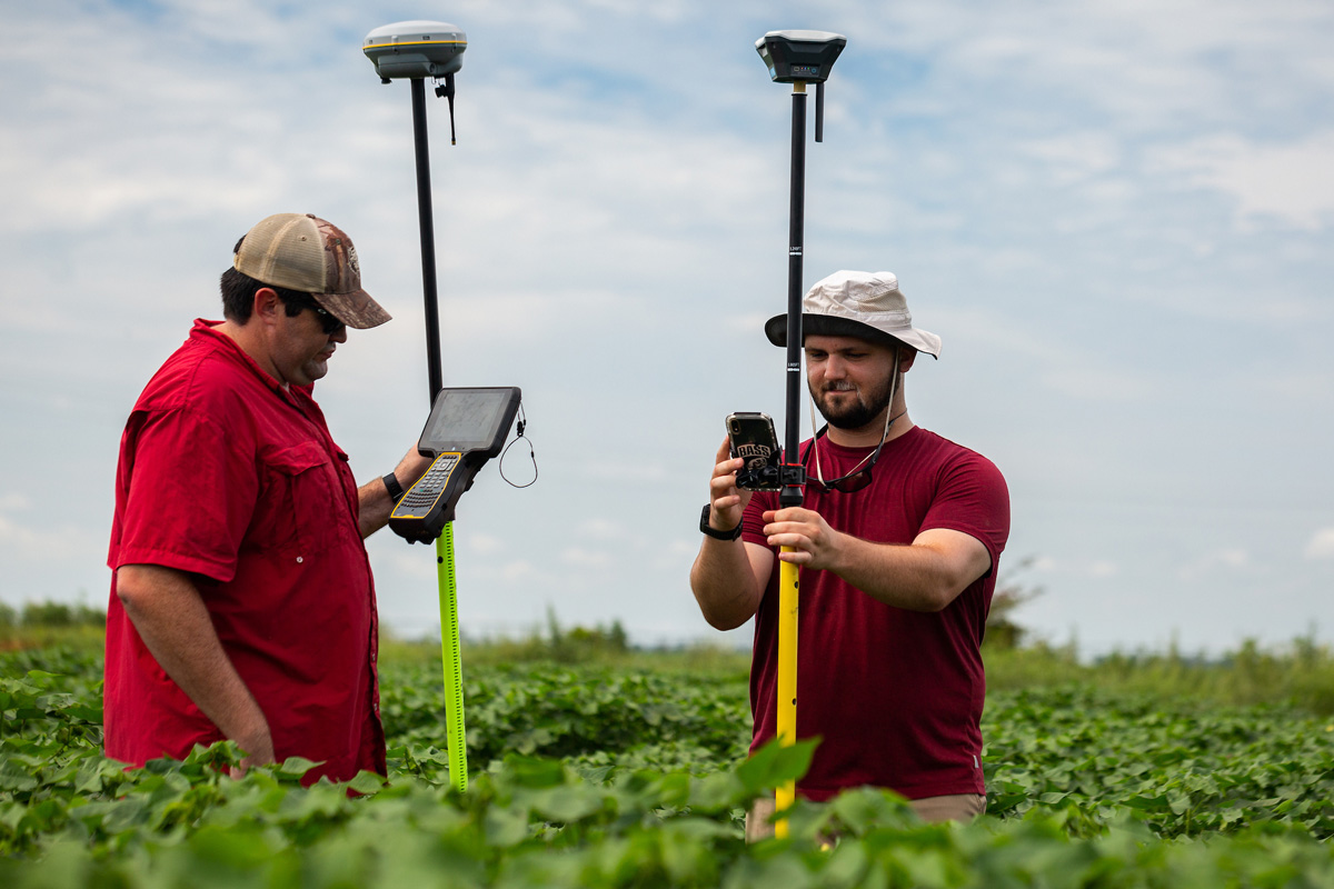 Two agriculture students use GPS surveying equipment in a green crop field.
