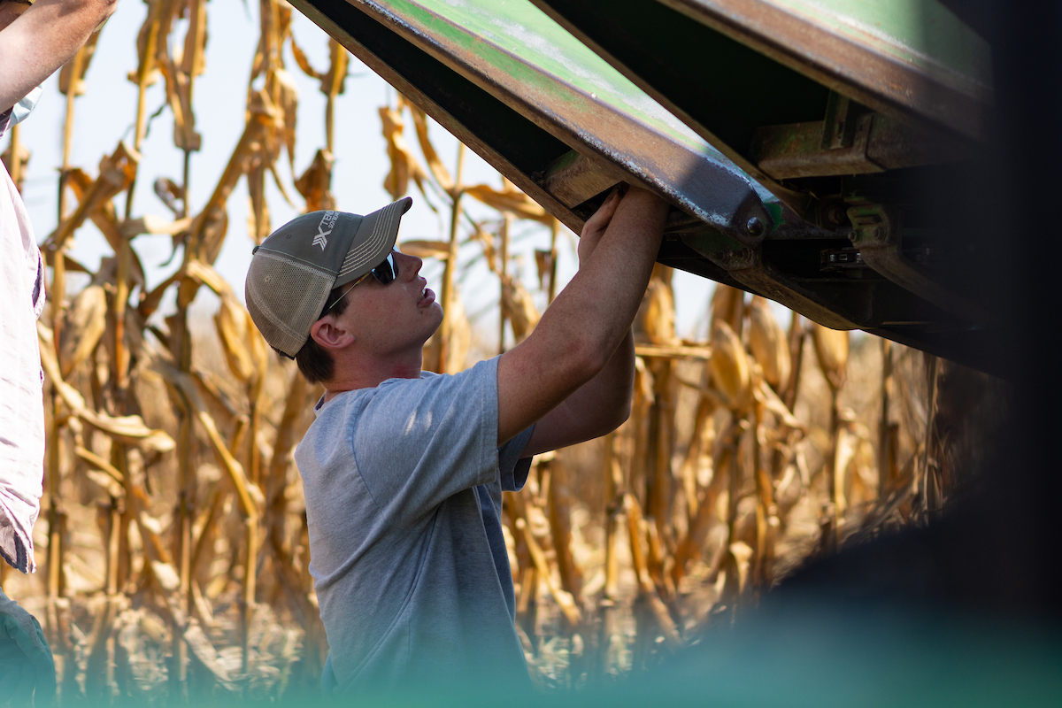 Student in cap and sunglasses works on farm equipment beside a cornfield.