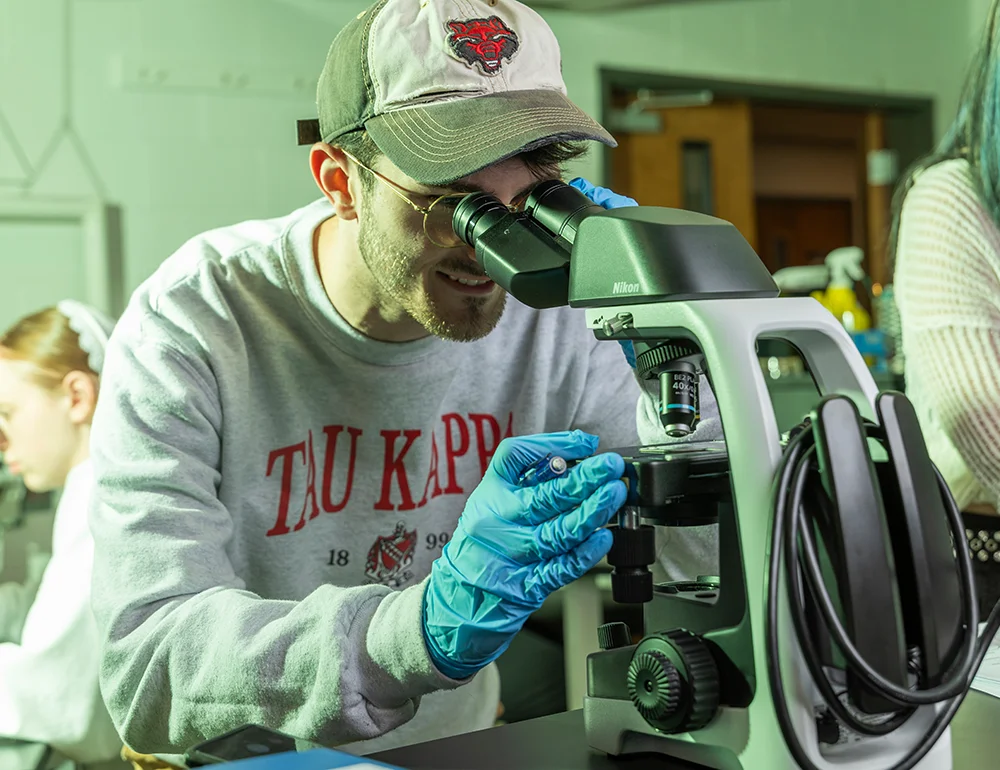 Student in a Red Wolves cap using a microscope.