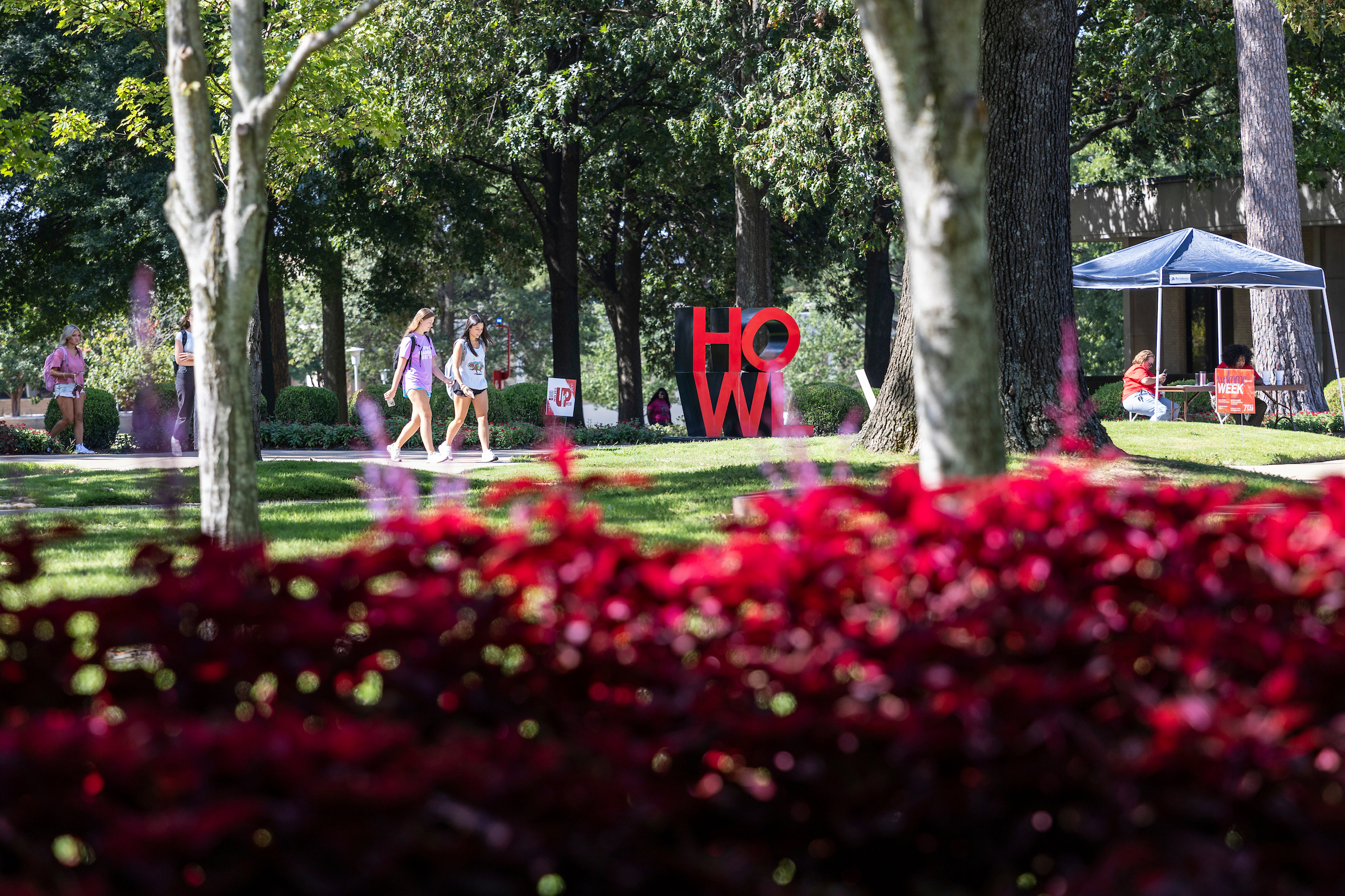 A-State students walk past the red HOWL sign with bright red flowers in the foreground on a sunny day.