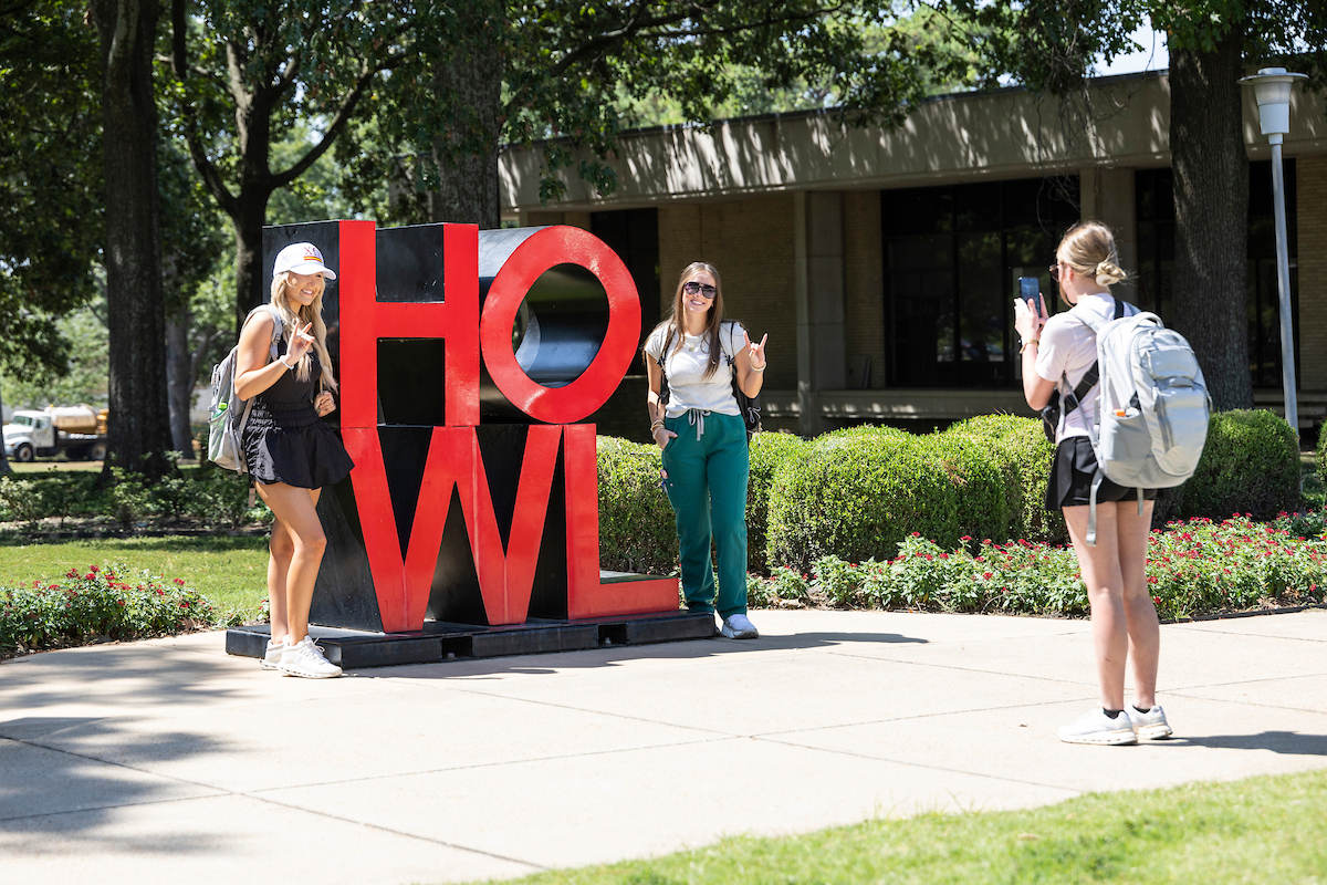 Two students pose with the red HOWL sign at ASU while a third takes their photo on a sunny day.