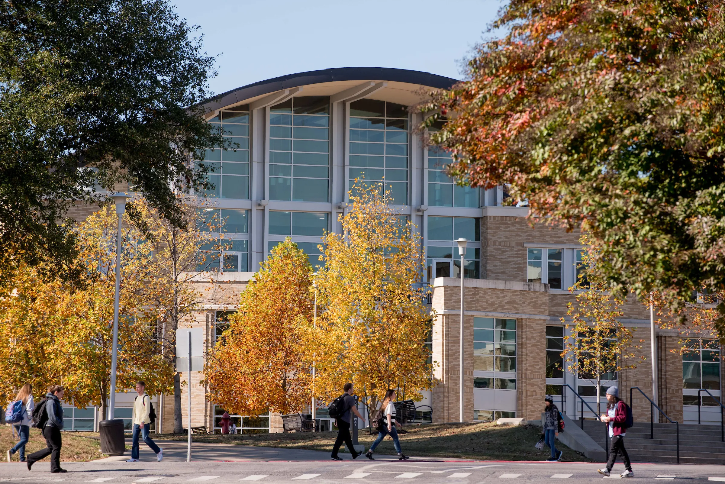 Students walk across campus near the Reng Student Union surrounded by golden autumn trees on a clear day.