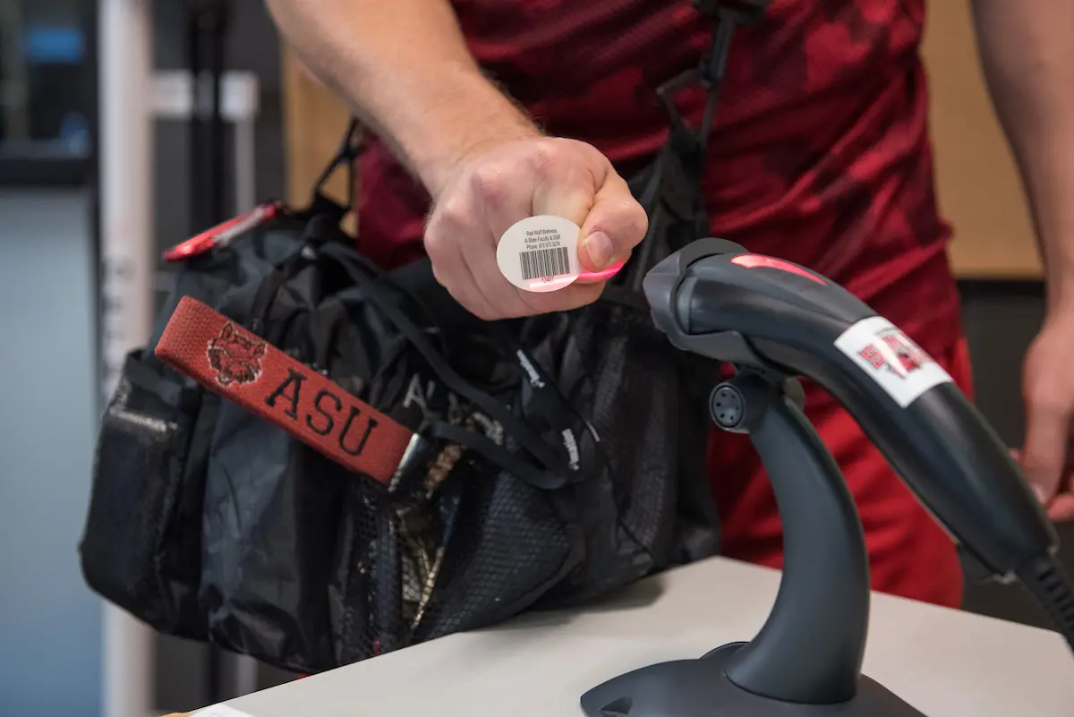 An employee scans a barcode with a handheld scanner at wellness check-in, wearing a bag with ASU Red Wolves lanyard.