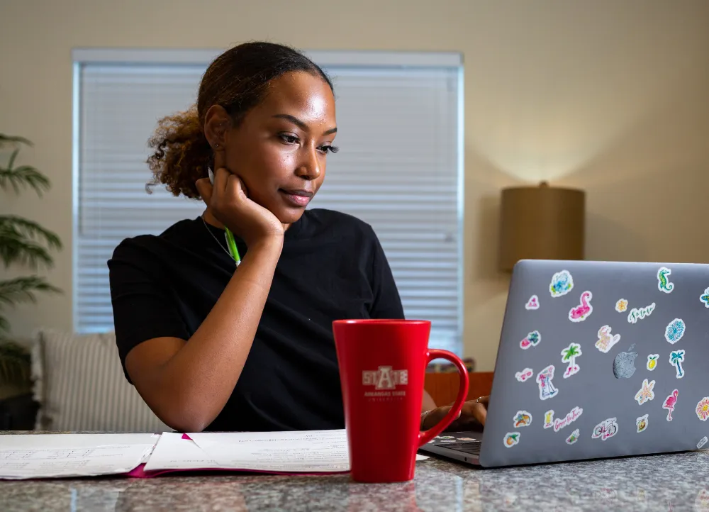 Student on laptop with A-State mug nearby.
