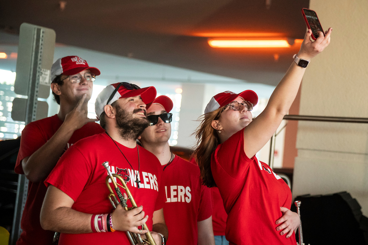ASU Howlers band members wearing red shirts take a group selfie while holding instruments in a parking garage.