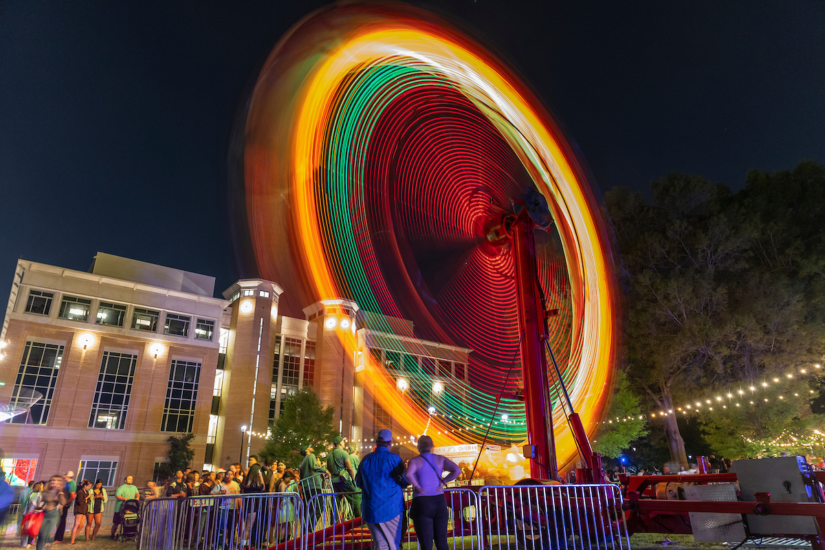 Nighttime long exposure of a colorful spinning ride at the campus “State Fair”, with crowds gathered near a brightly lit building.