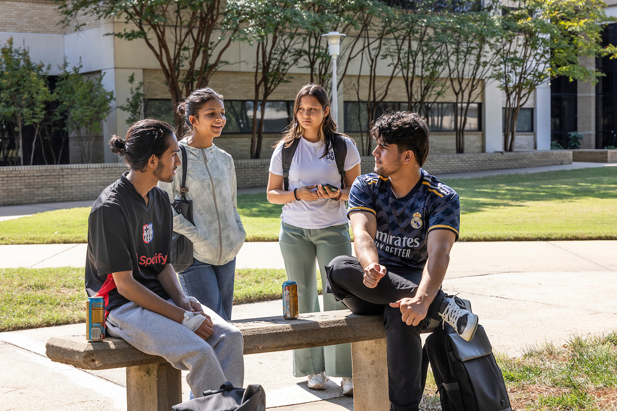 A-State students gather around a bench chatting in the shade on a sunny day near campus buildings.