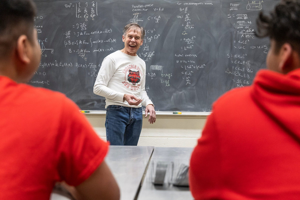 Physics professor teaches students with chalkboard full of equations in background.
