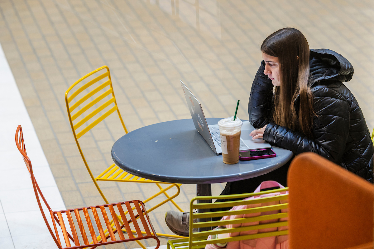Student in a black jacket studies with a laptop and iced coffee.