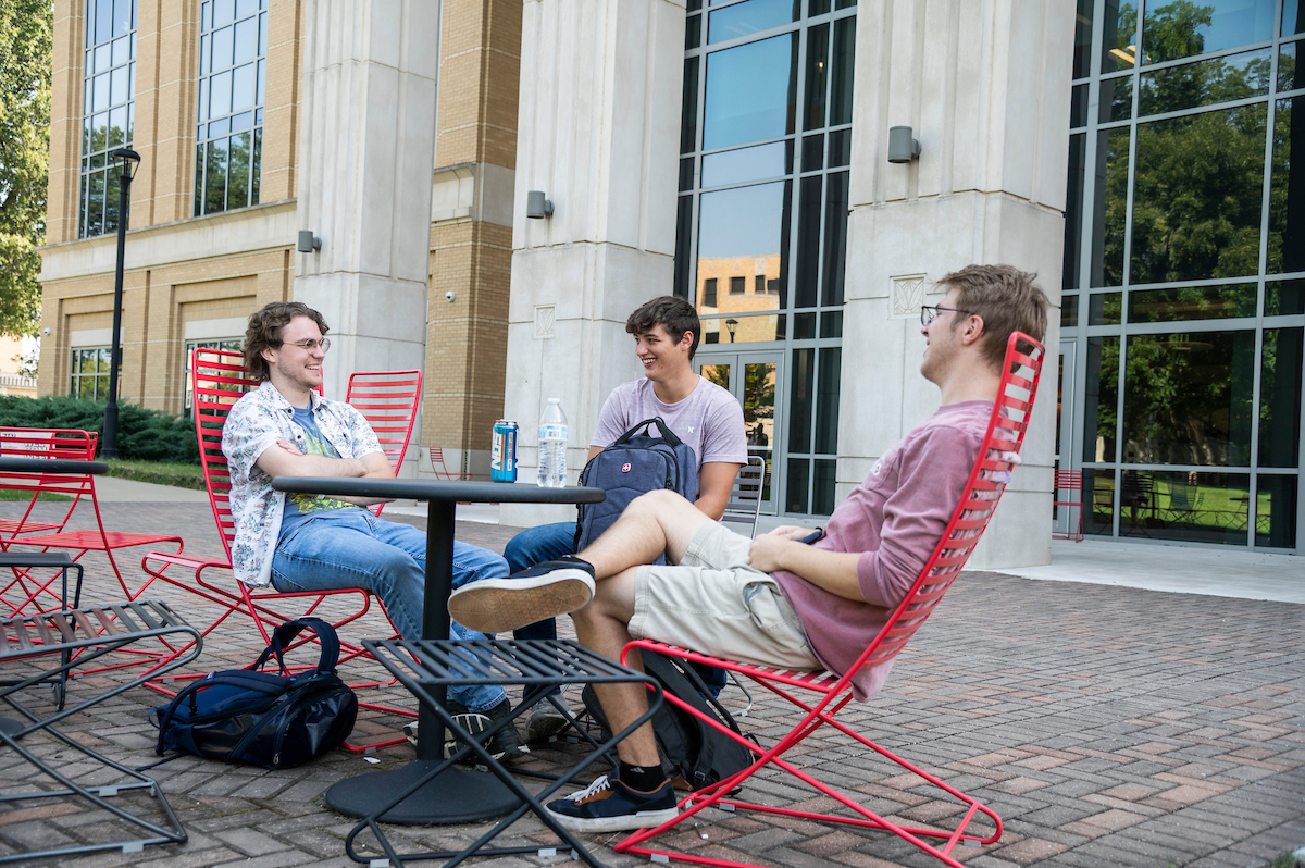 Three A-State students sit and chat outside the campus library on red chairs around a table on a sunny day.
