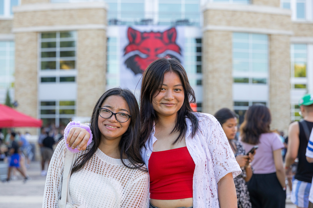 Two A-State students smile in front of a Red Wolves banner during a vibrant campus event.