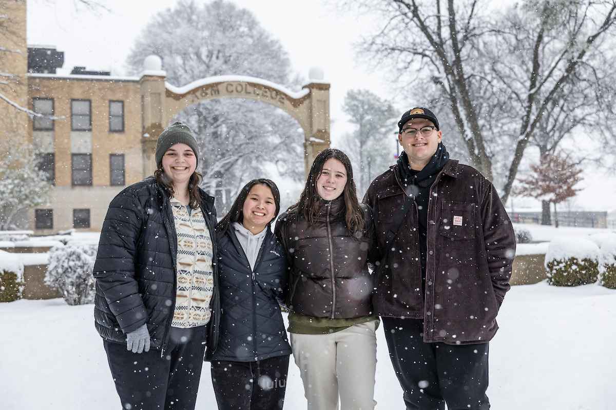 Four students pose together in falling snow near the historic A&M College arch on campus.