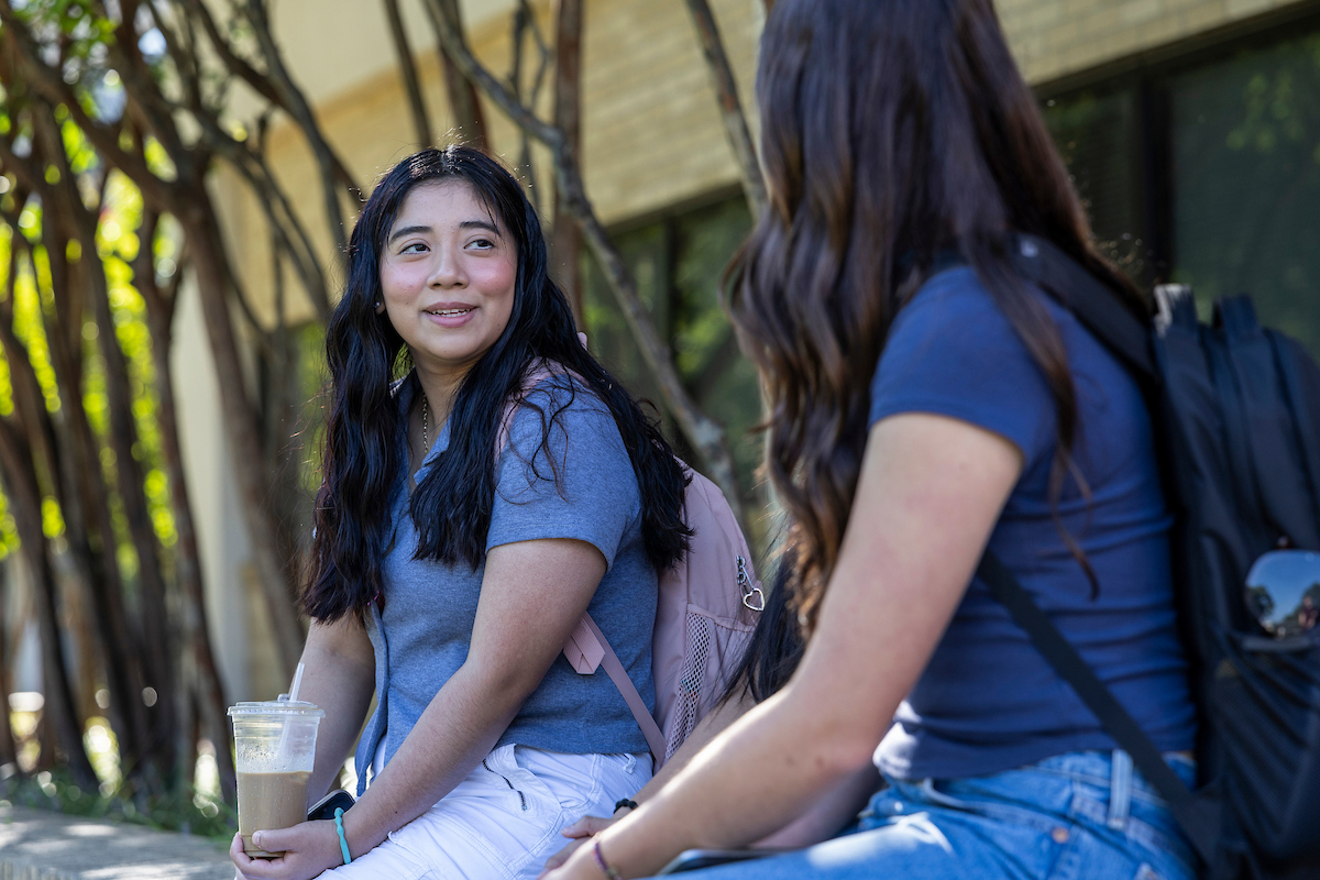 A-State students talk and relax on a bench outside on a bright sunny day.