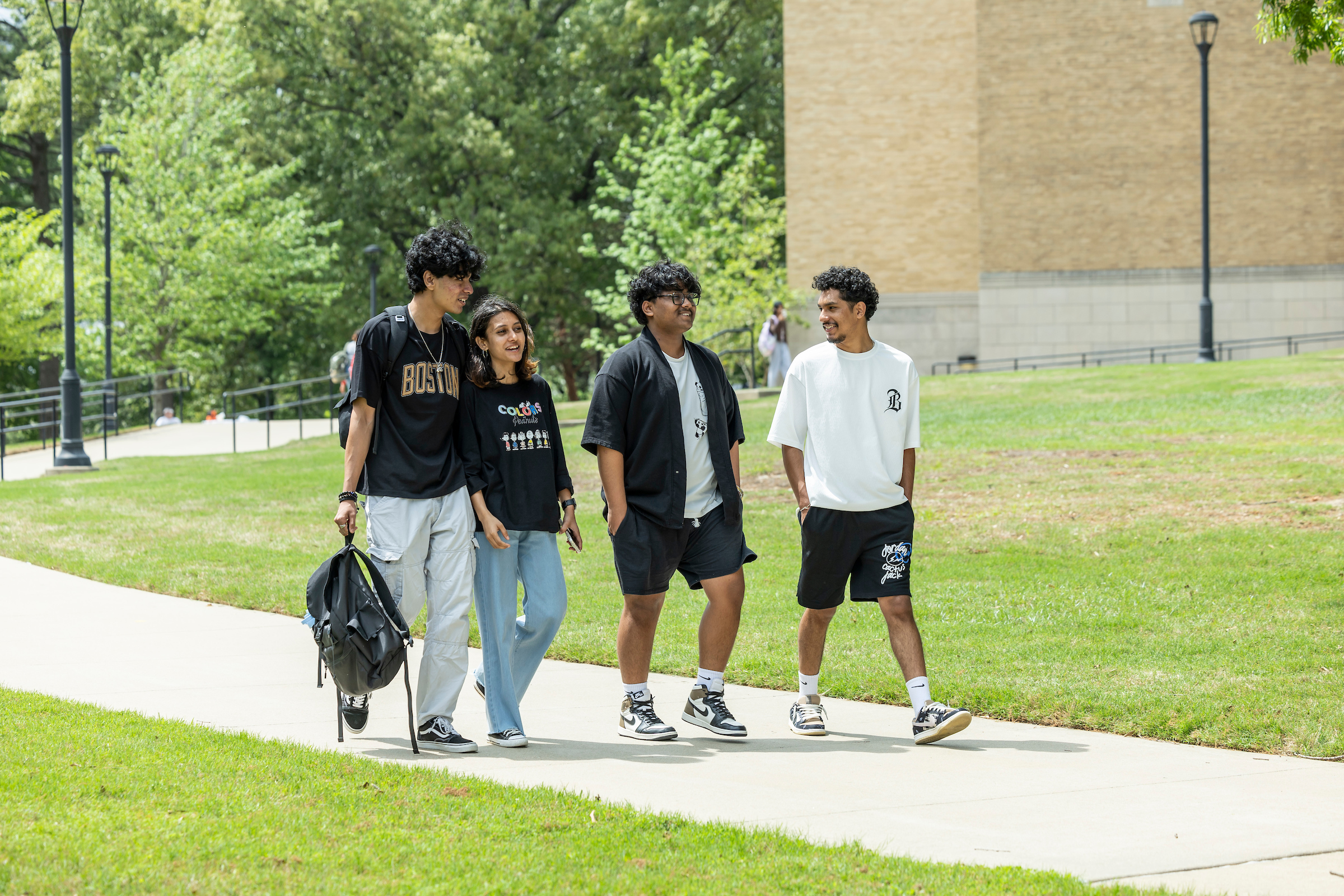 Group of A-State students walk and talk along a campus sidewalk on a sunny green day.