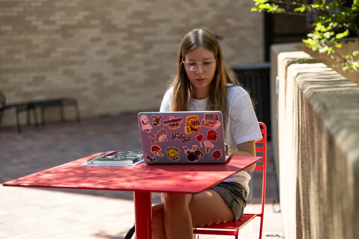 A-State student works on a colorful sticker-covered laptop at a red outdoor table.