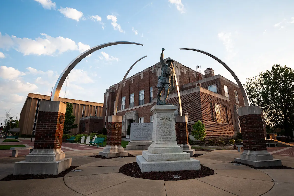 Craighead County Veterans Memorial features a bronze soldier statue under arching metal beams at sunset near the courthouse.