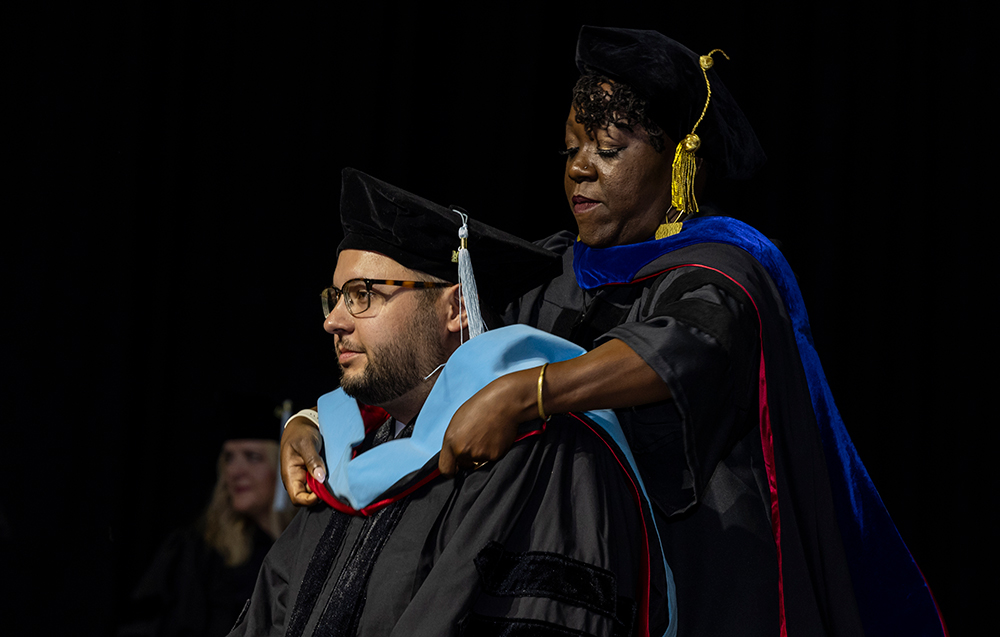 Faculty member places doctoral hood on graduate during A-State summer commencement ceremony on a dark stage.