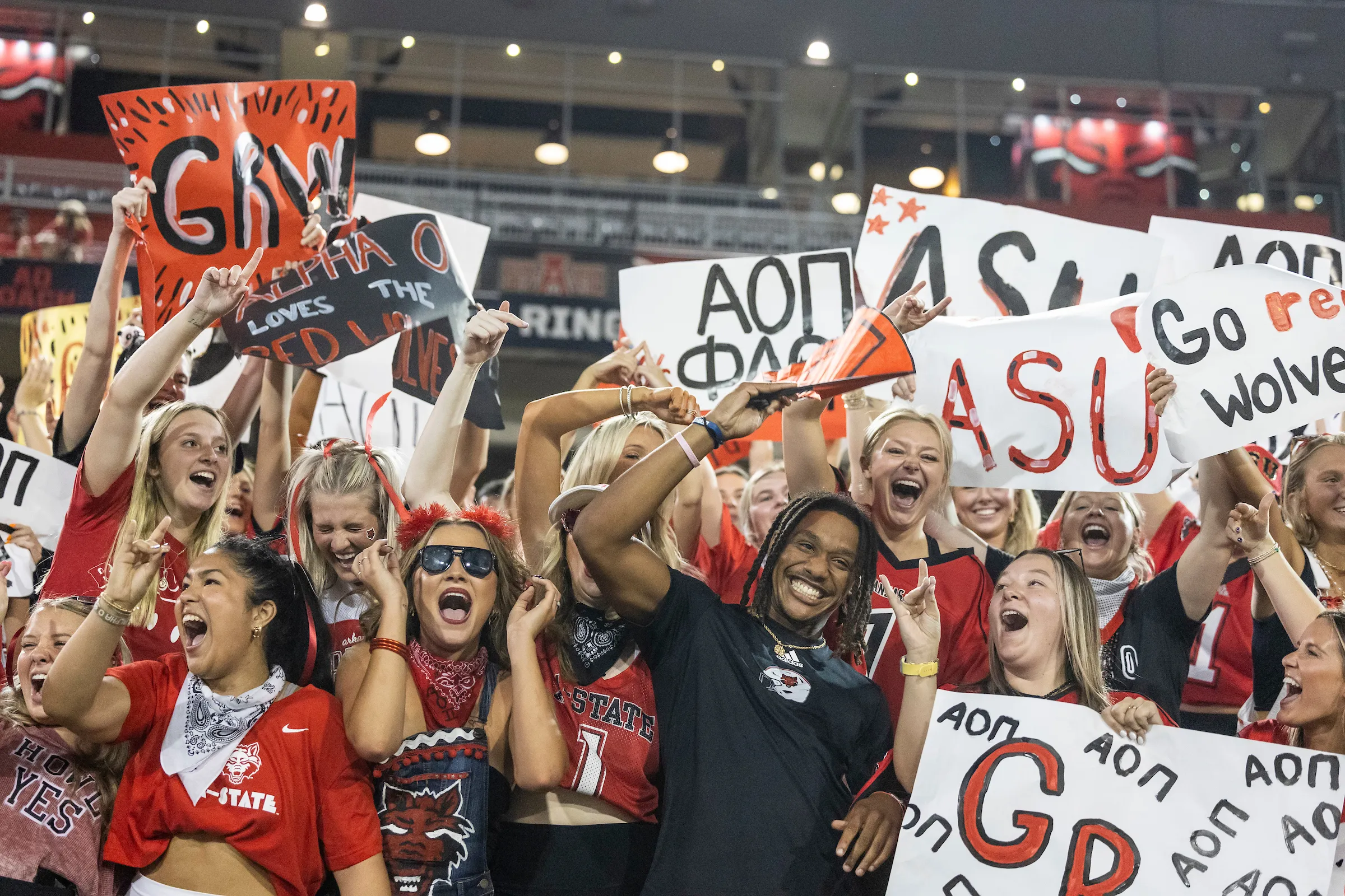 Energetic crowd of Greek life students cheering with custom signs at an A-State football game.