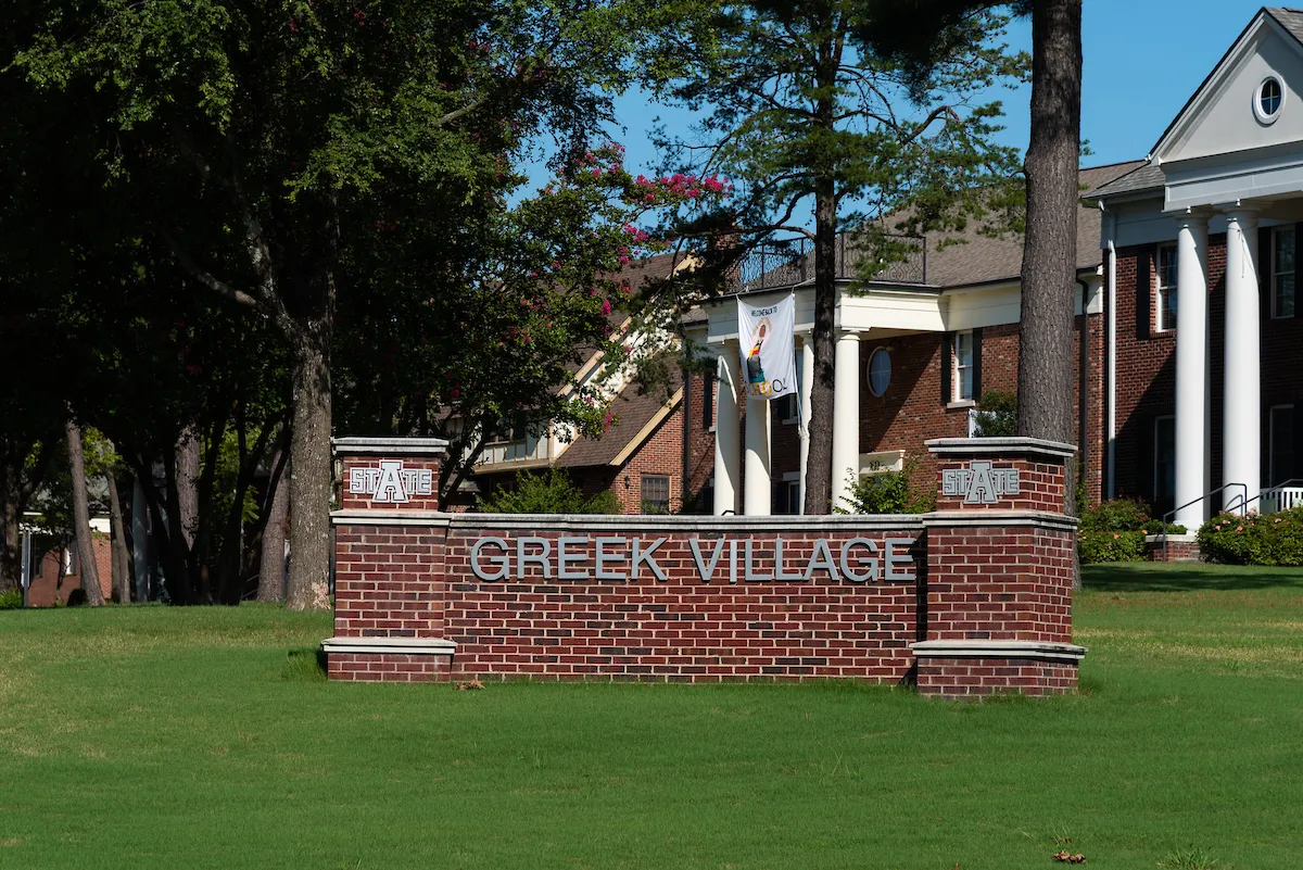 Brick Greek Village sign at A-State with fraternity houses and trees in the background.