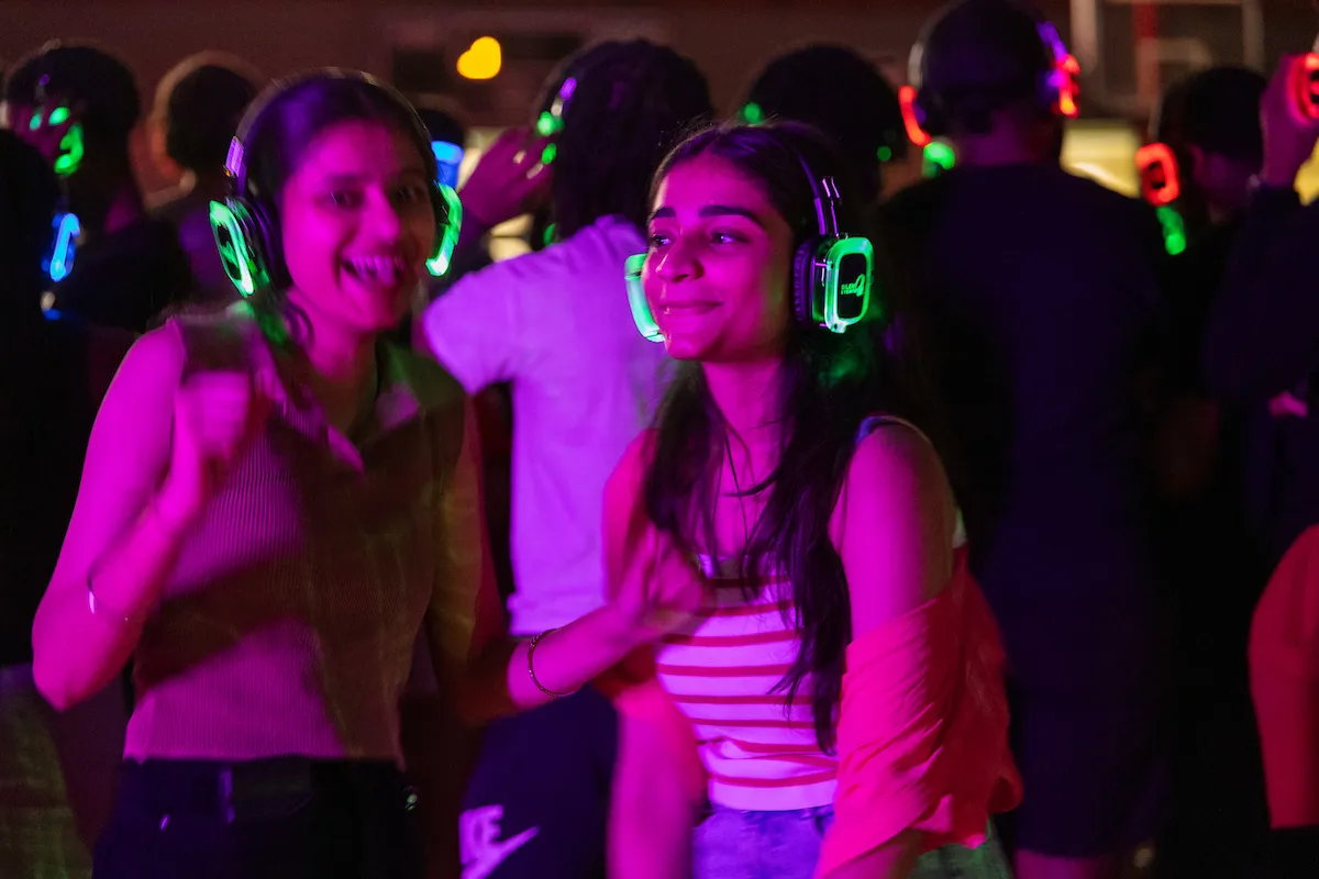 Two students dance and smile with glowing headphones at a silent disco party with purple lighting.