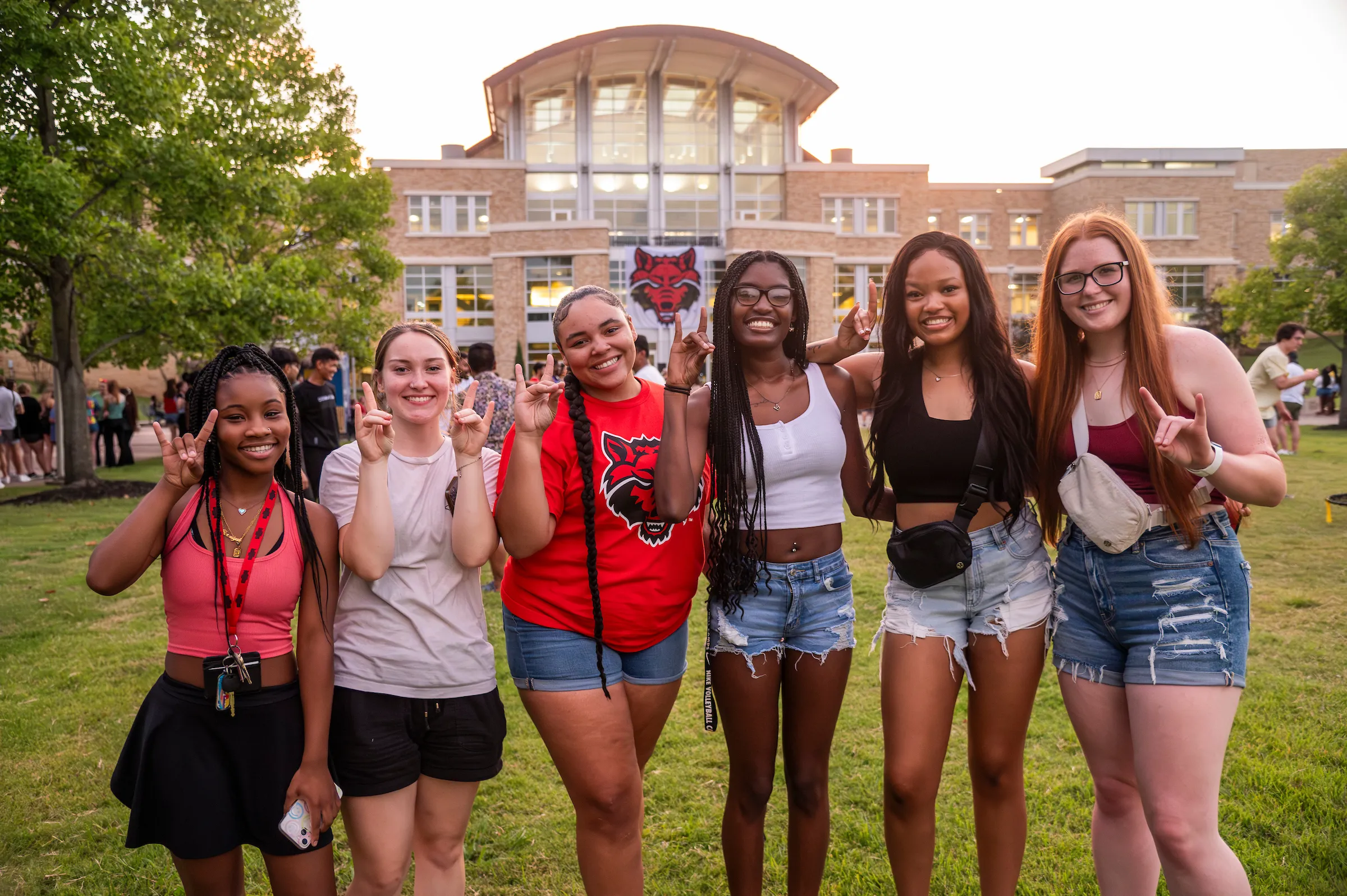 Group of six smiling students flash wolf hand signs in front of the Student Union during a campus evening event.