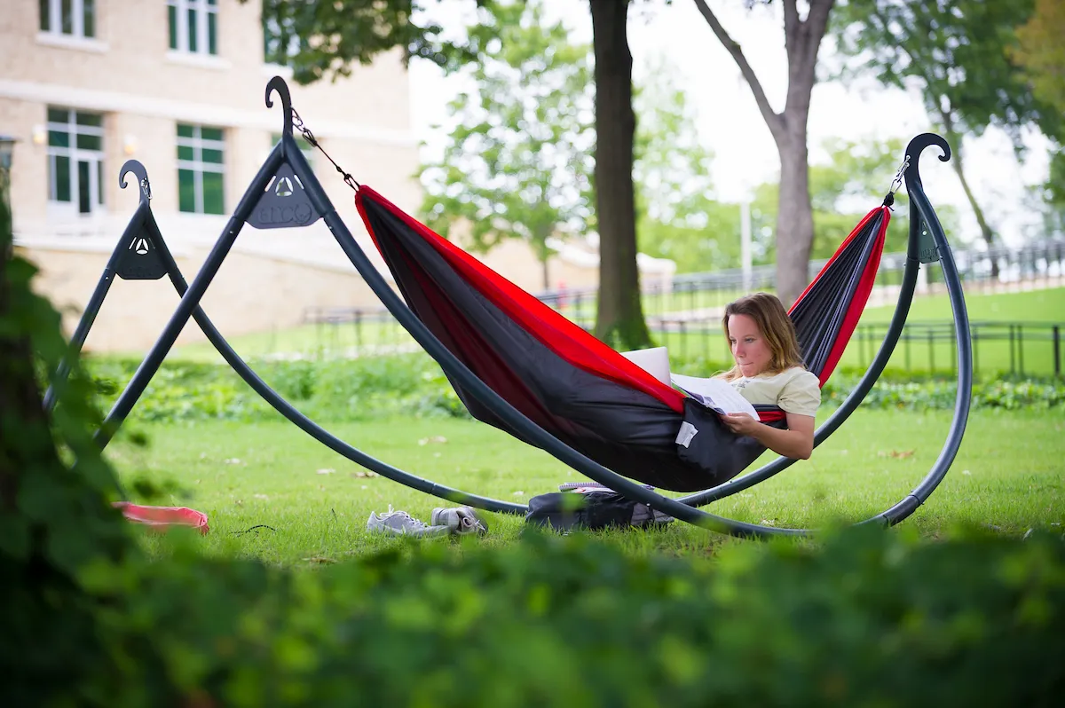 Student relaxes in a red-and-black hammock studying with laptop and papers on a grassy campus lawn.