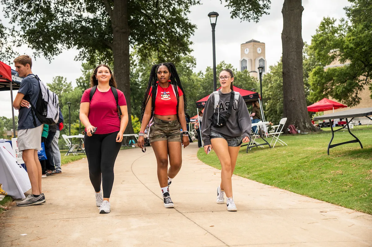 Three students walk a sidewalk lined with booths and tents during a campus event with the bell tower in background.