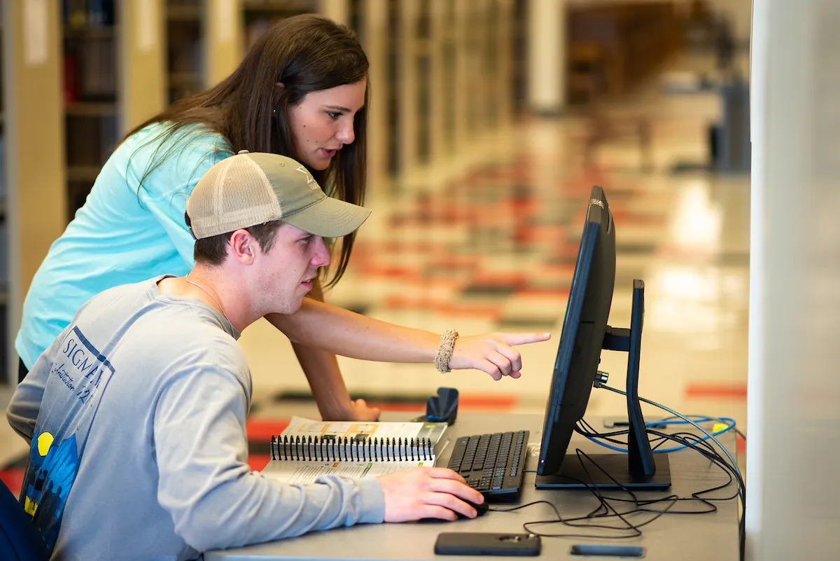 Female student points to a screen while helping a classmate on a desktop computer in a library setting.