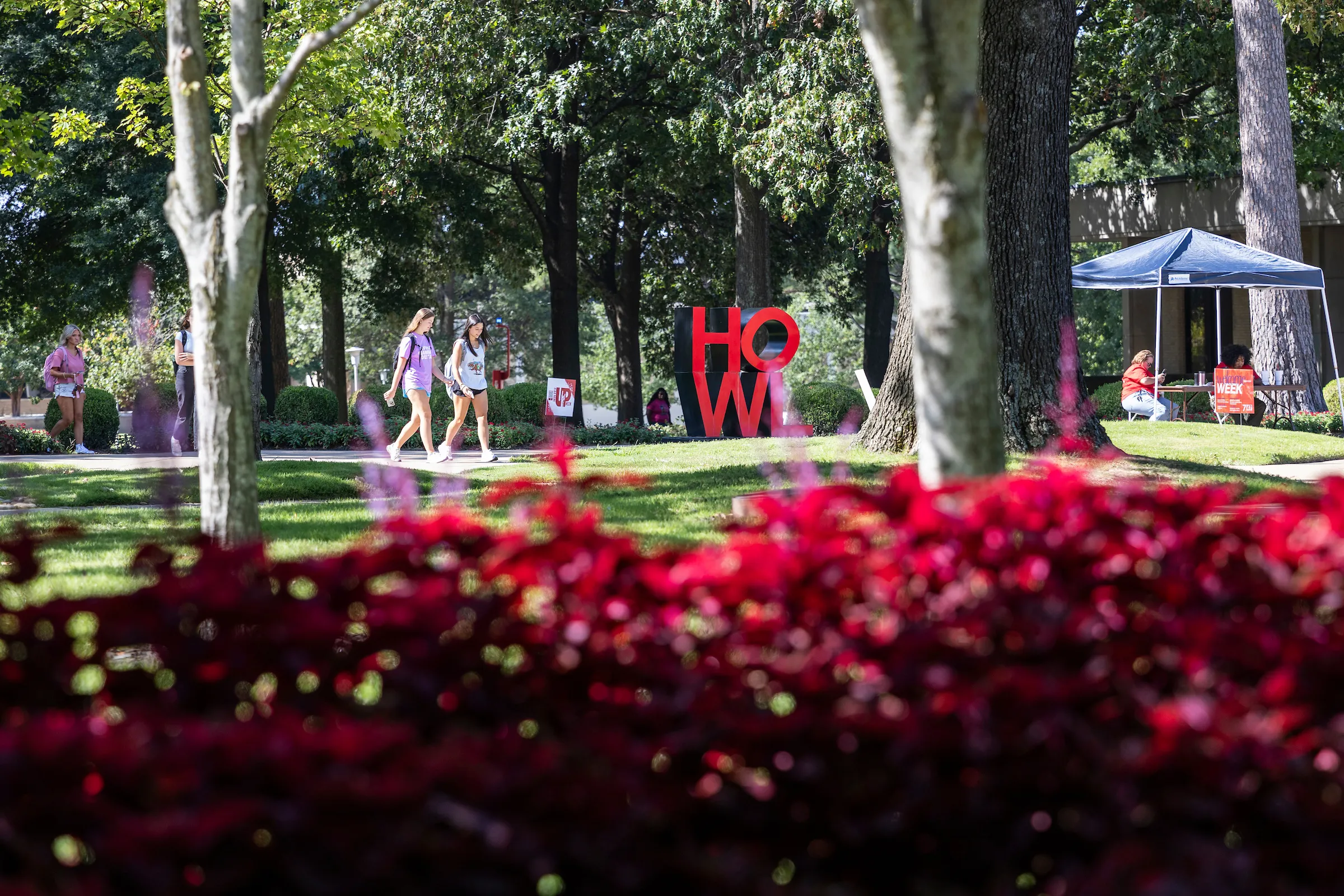 Students walk past red flowers and trees near the HOWL sculpture on a vibrant first day of classes.