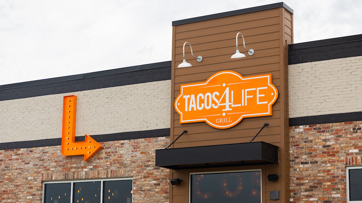 Bright orange “Tacos 4 Life Grill” sign on a brick building with vintage arrow decoration.