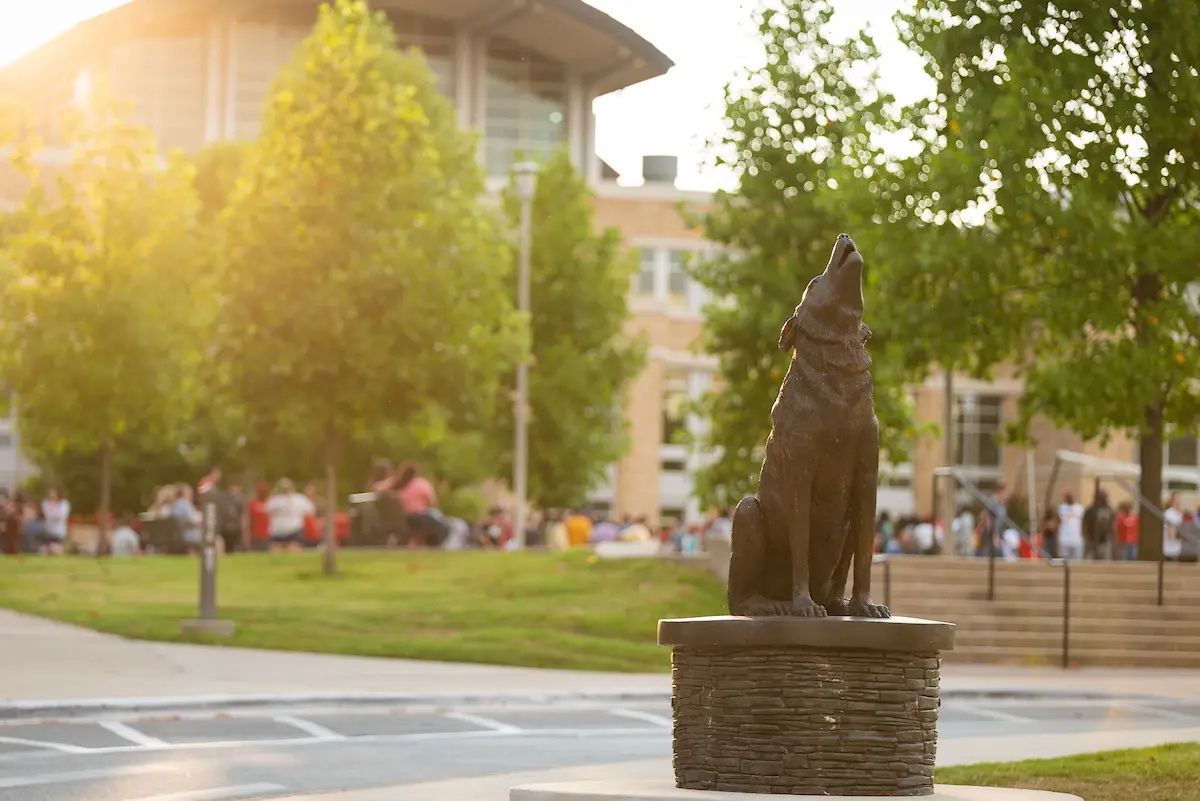 Bronze howling wolf statue on the A-State campus at sunset with blurred students and trees in the background.