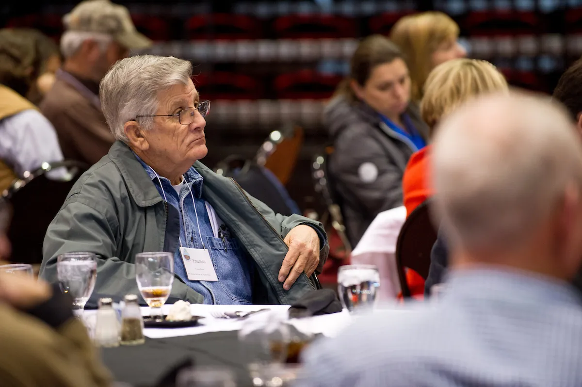 Older man in glasses listens attentively during a conference session at a round table.